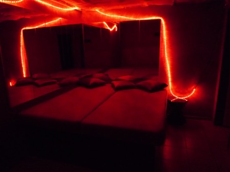 Spiegelzimmer mit großer Liege, Kissen und roten Lichterketten an Spiegeln und Decke.